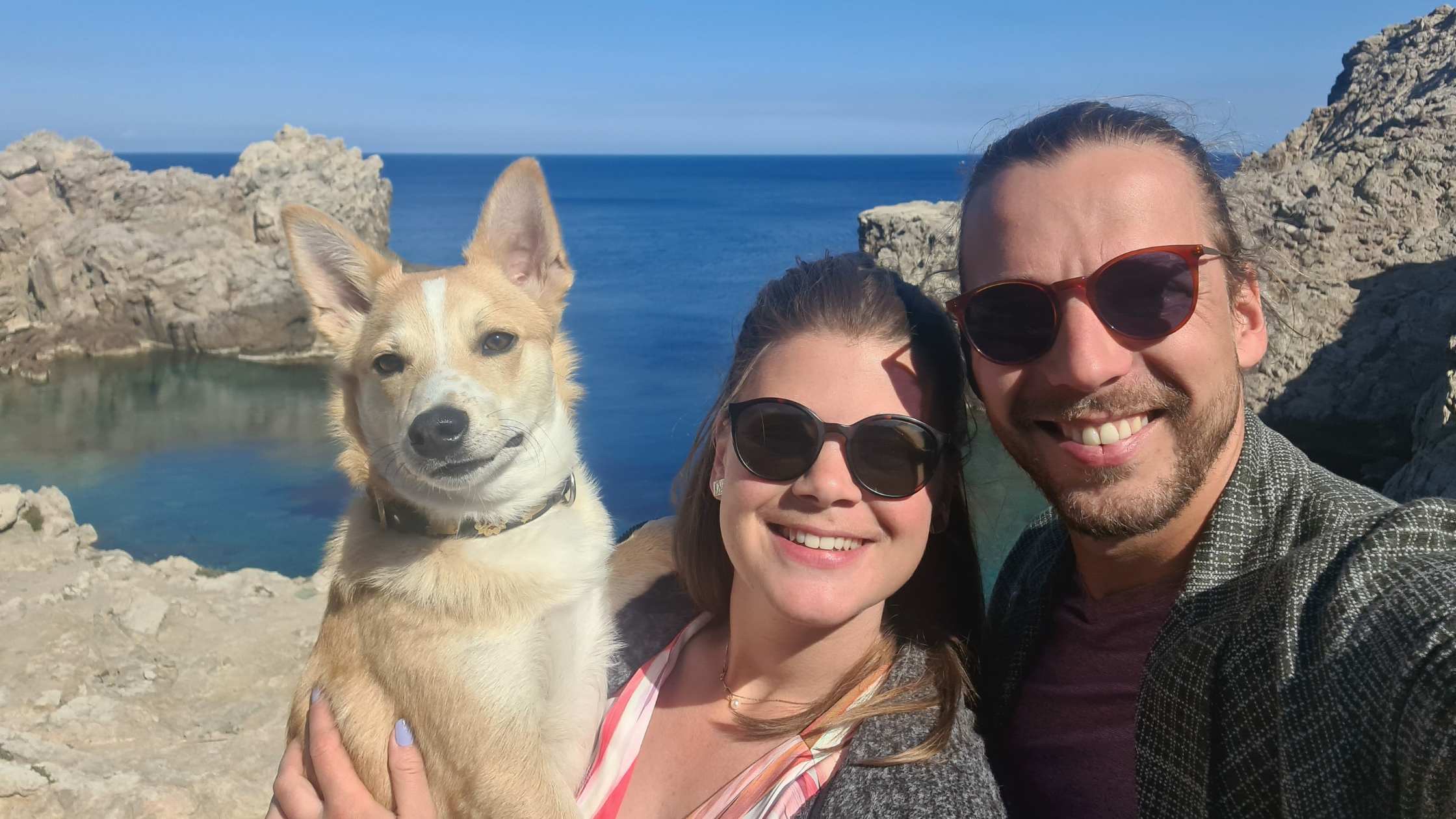 Anne, Pascal and their dog Shiraz are all facing the camera with the blue sea and some rocks behind them