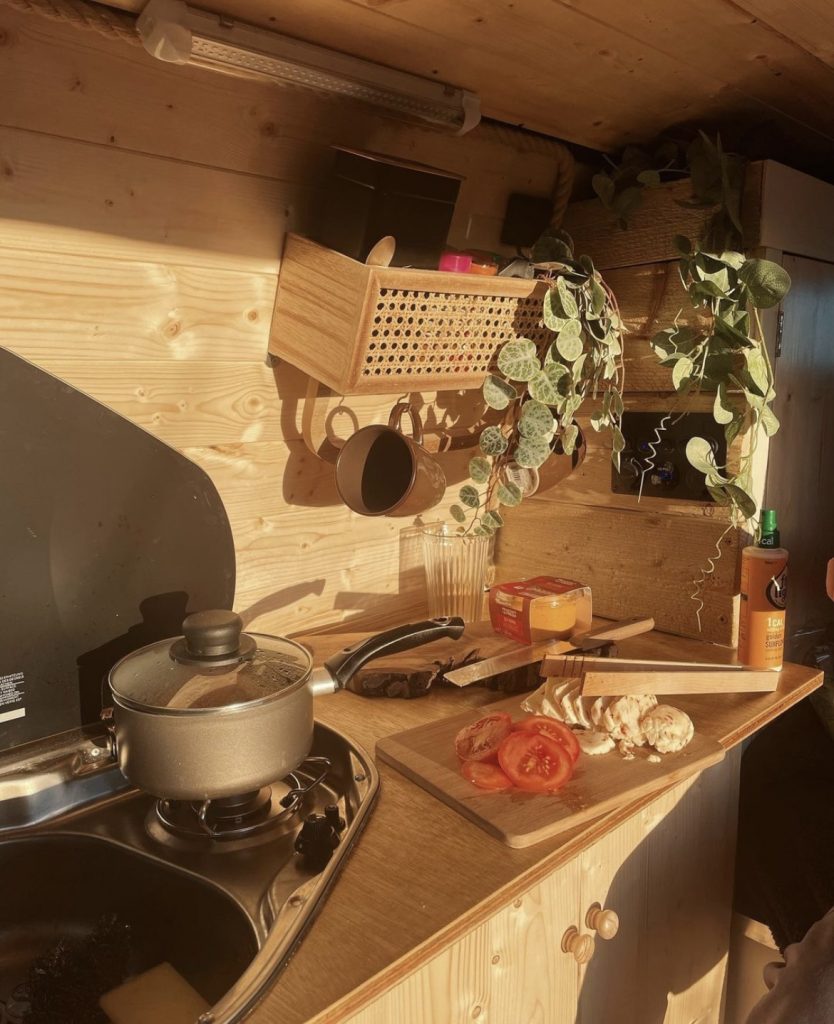 Inside the campervan kitchen. The walls, cupboard, and worktop are all made of light wood. There is a saucepan on a single hob and chopped fresh tomato and cheese on a chopping board.