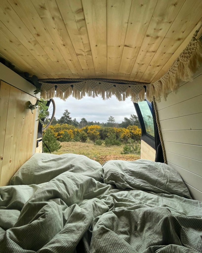 The view is from the end of the bed in the campervan looking out the open back doors. The bedding is a little crumpled. There is macrame bunting & the view is of lots of greenery.