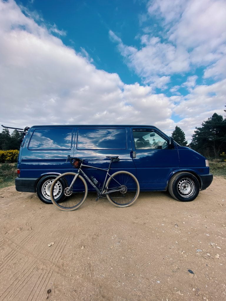 Blue T4 camoervan parked up on a dirt track. The view is side on on the drivers side and there is a bicycle leaning up against the side.
