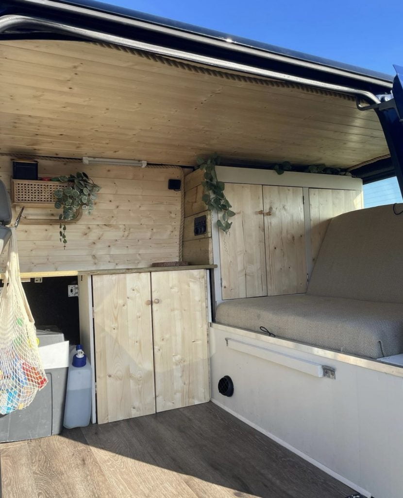 The inside of the campervan, there is light wood cladding throughout with a beige seat. There is a mesh shopping bag hanging up and green plants trailing down from the shelves.