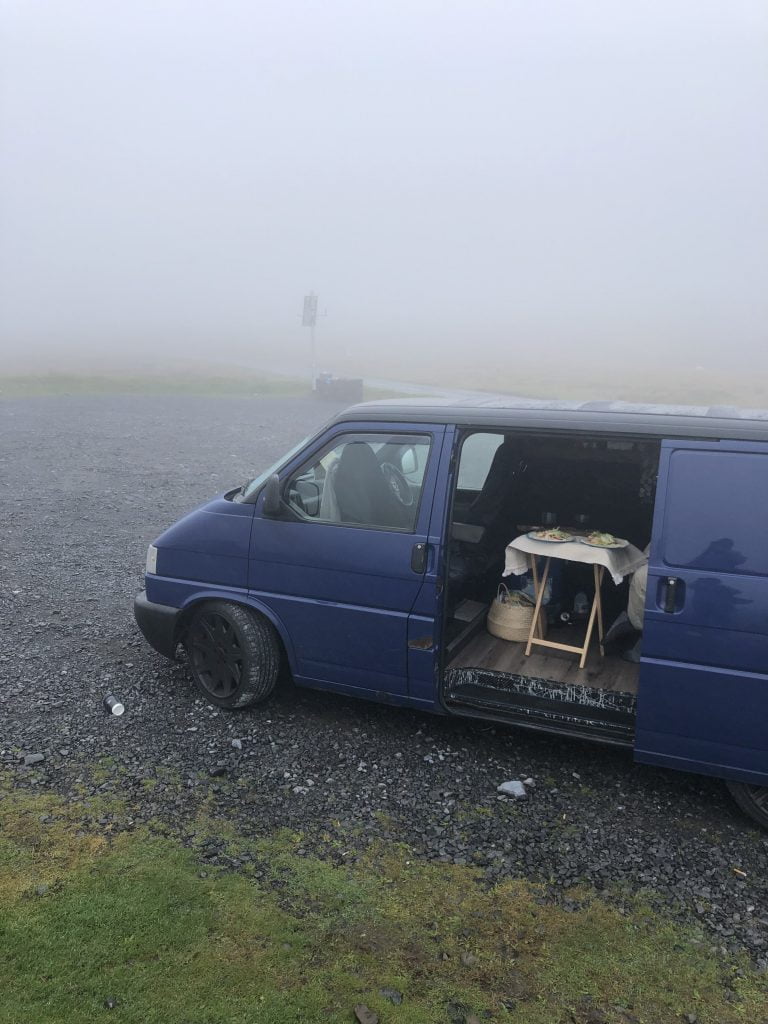 A blue T4 campervan is parked up surrounded by fog. The sliding door is open & there is a table set for 2 with 2 plates of food.