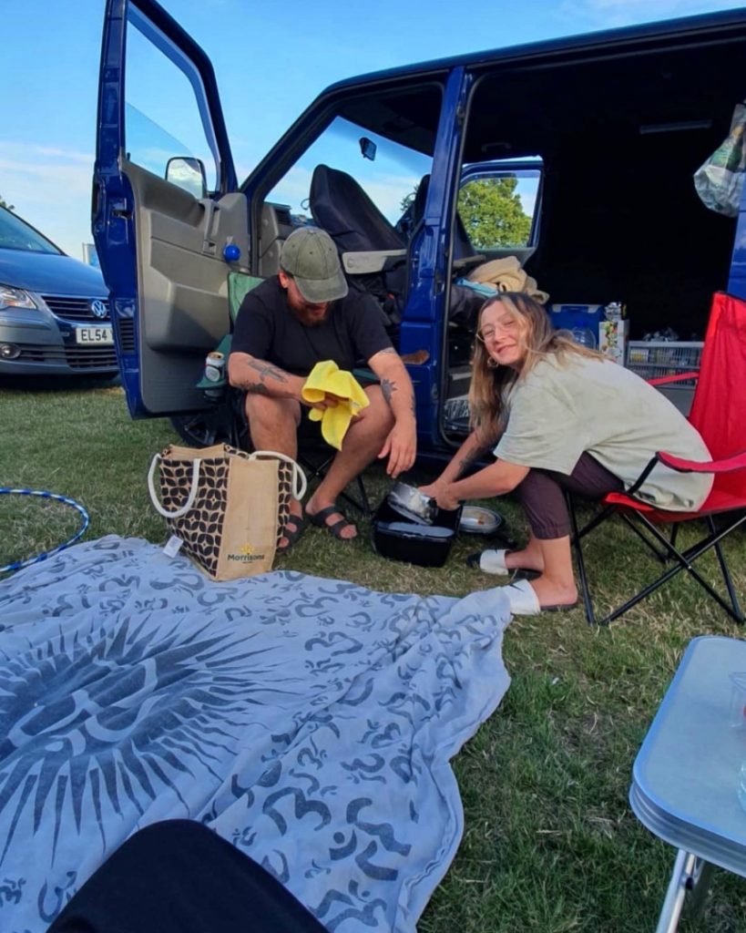 The couple are sat washing up their pans outside the van. Leanne is looking at the camera smiling, whilst washing a saucepan in a small black washing up bowl. Billy is sat on a camping chair with a tea towel in his hand, looking down.