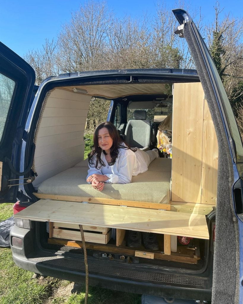 Leanne is dressed in white, she is lying on the bed on her front. The back doors of the campervan are open and the sun is shining, Leanne is smiling.