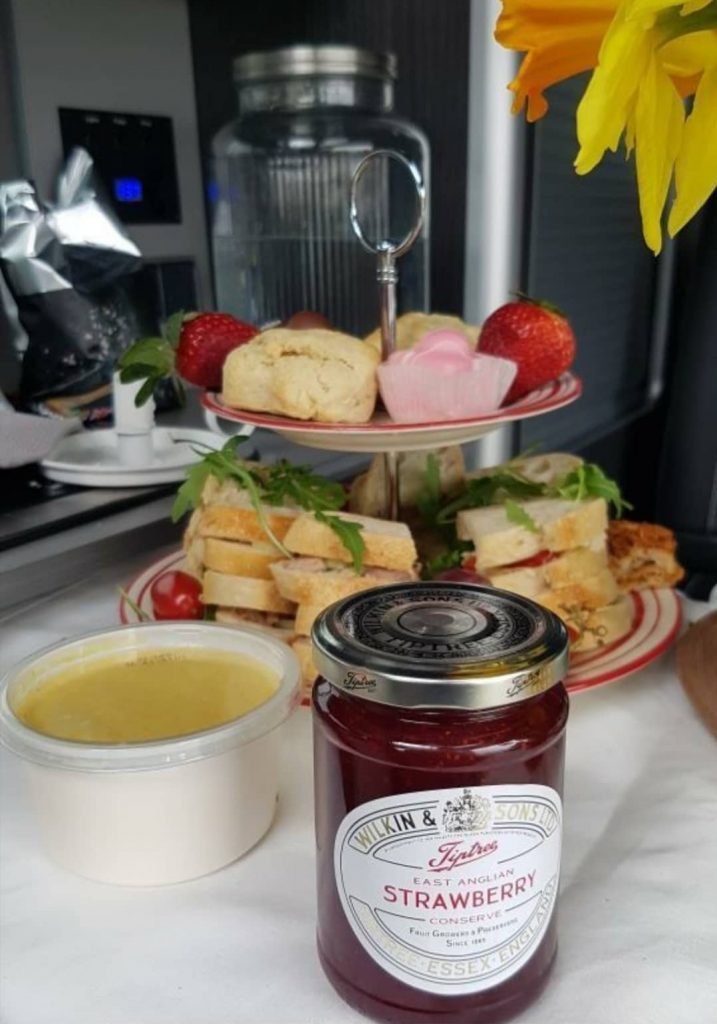 Afternoon tea presented on a red and white cake stand. On the top plate there are 2 scones and 2 french fancies as well as 2 straweberries. On the bottom plate are some sandwiches with salad