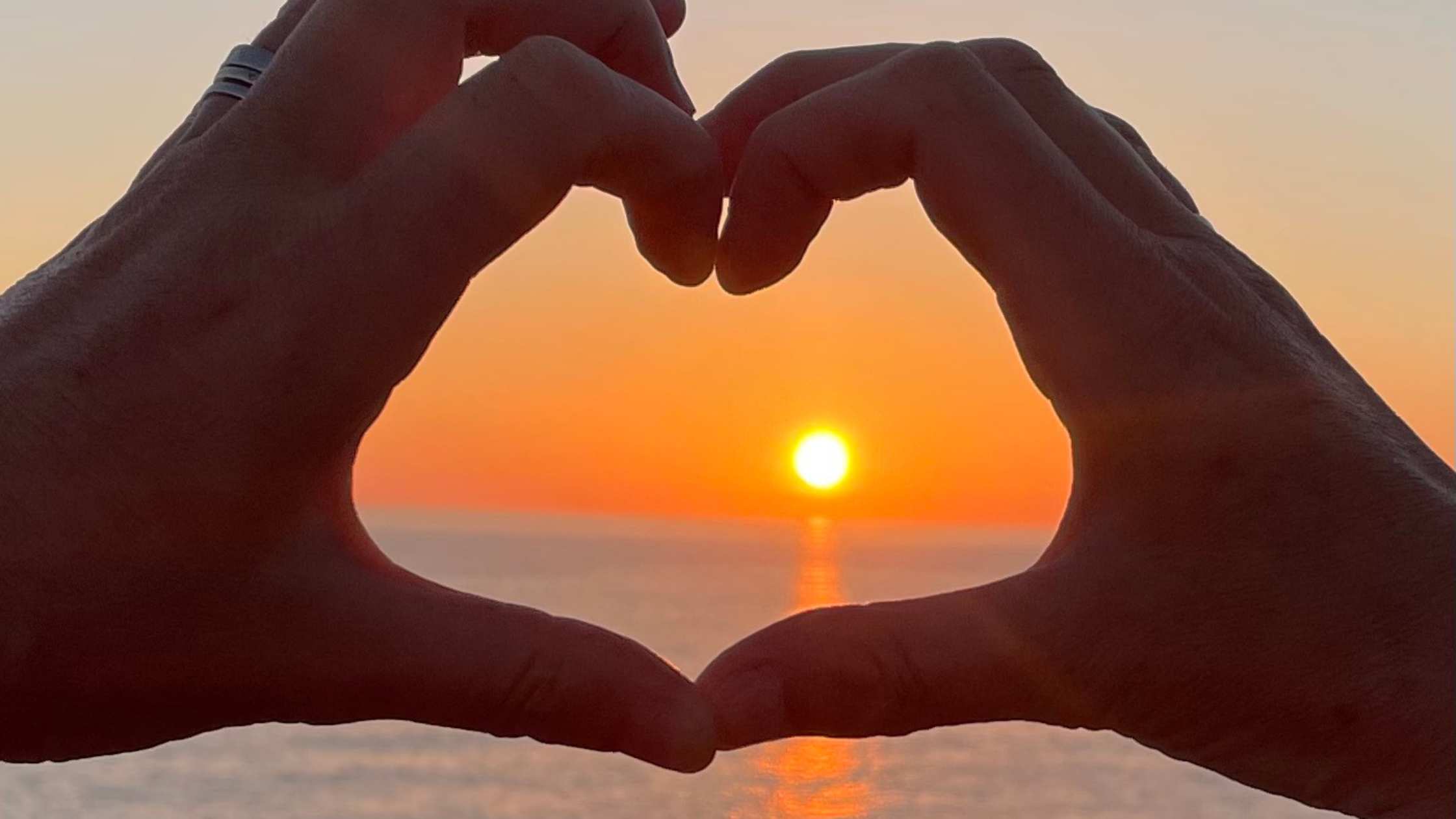 2 hands joined together in the shape of a heart in front of a sunset over the sea.